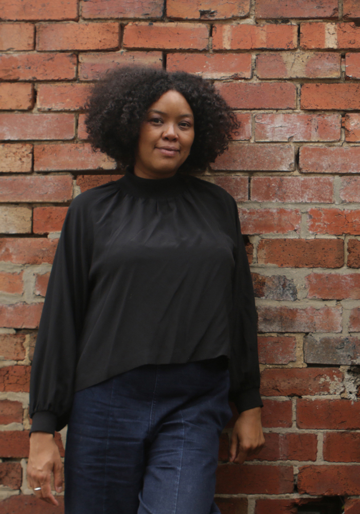 An African woman with big curly hair wearing a black long sleeve top and dark denim jeans smiles quietly in front of a red brick wall.