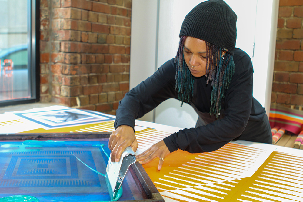 An Afro-Carribean woman with blue and purple dreadlocks is busy screen printing and looking down at her work with great focus. She is holding a squeegy moving teal colour paint ink above a square screen print frame, across a blue colour exposure film, situated on a table with an orange and white stripe-like pattern. She is wearing a black long sleeve top, black apron, and a black beanie leaning across the table, in front of two windows surrounded by brick walls. 