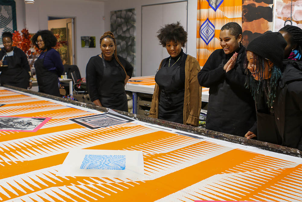 Seven African women are standing along a long table that has a print with orange and white stripe-like repeating pattern. There are black and white images on the table, adjacent to two diamonds, one with a pink border and black and white image of an African woman inside and the one in the foreground has no border, and a blue printed image of a woven material. Two African women on the right are looking down at the table amazed with big smiles, as the others standing beside the table are looking on and at the long print. In the background is the studio with hanging prints on the white walls, and two doors, one closed, another open with light emitting from another room.