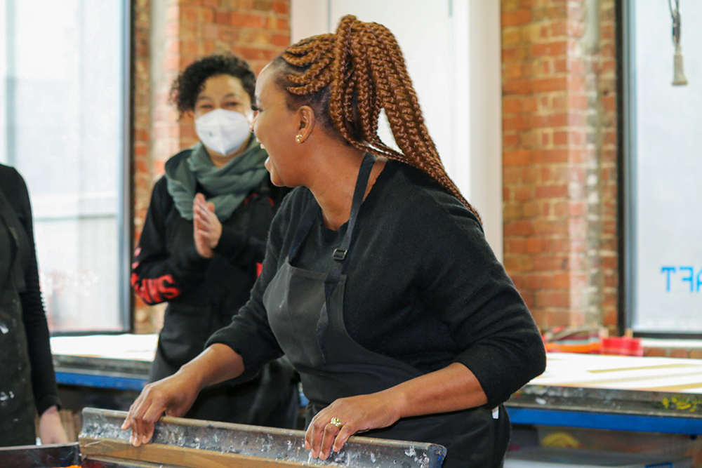 An African woman with copper colour thick braids tied into a pony tail, wearing all black and a black apron, holds a squeegy, is laughing looking to the left. An African woman in the background with curly short hair, wearin a green scarf and black clothes, with a white respitory mask is clapping with eyes smiling. They are inside brick studio with large windows and a long table in the background.