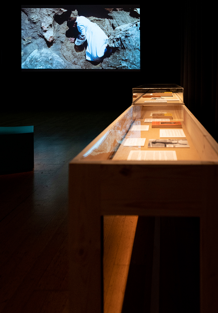 A dark room has a vitrine table in the foreground housing a collection of photos, texts and books, and a film projection of an African woman wearing all white is lying within a rocky environment on the screen.