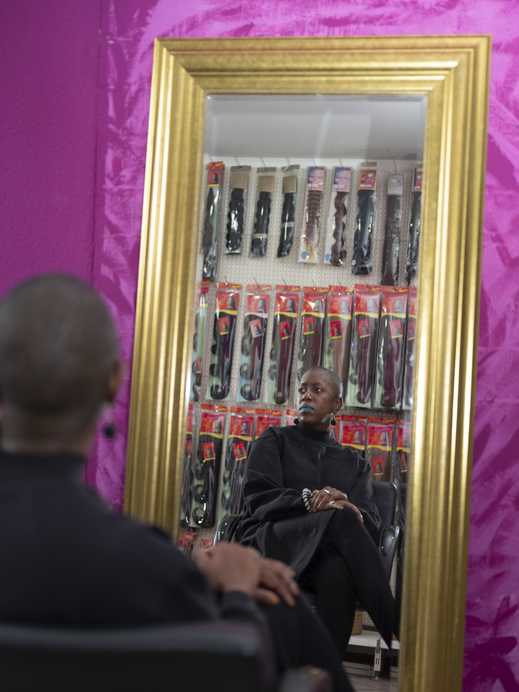 A pink room shows the back of a black woman with a shaved head, with her reflection visibale within a gold frame mirror. She has blue lipstick sitting crossed legged looking left, within a backdrop many packets of African hair extensions. 
