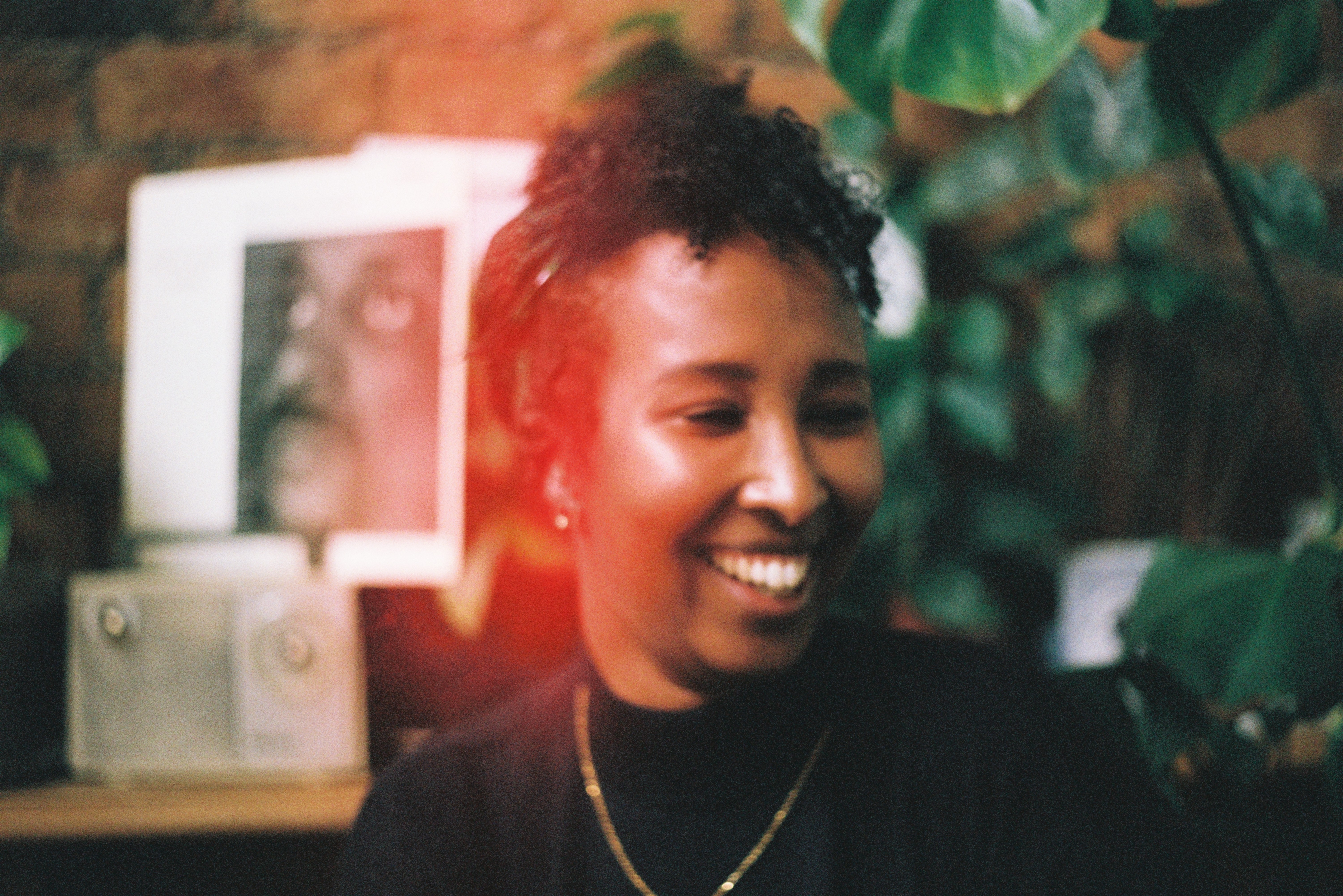 An African woman wearing a black top with a gold chain necklace is laughing looking to the right, with short curly hair tied up, sitting in a room with green leafed plants in the background that is out of focus. Behind her on the left and vinyl record sleeves and an old radio. 