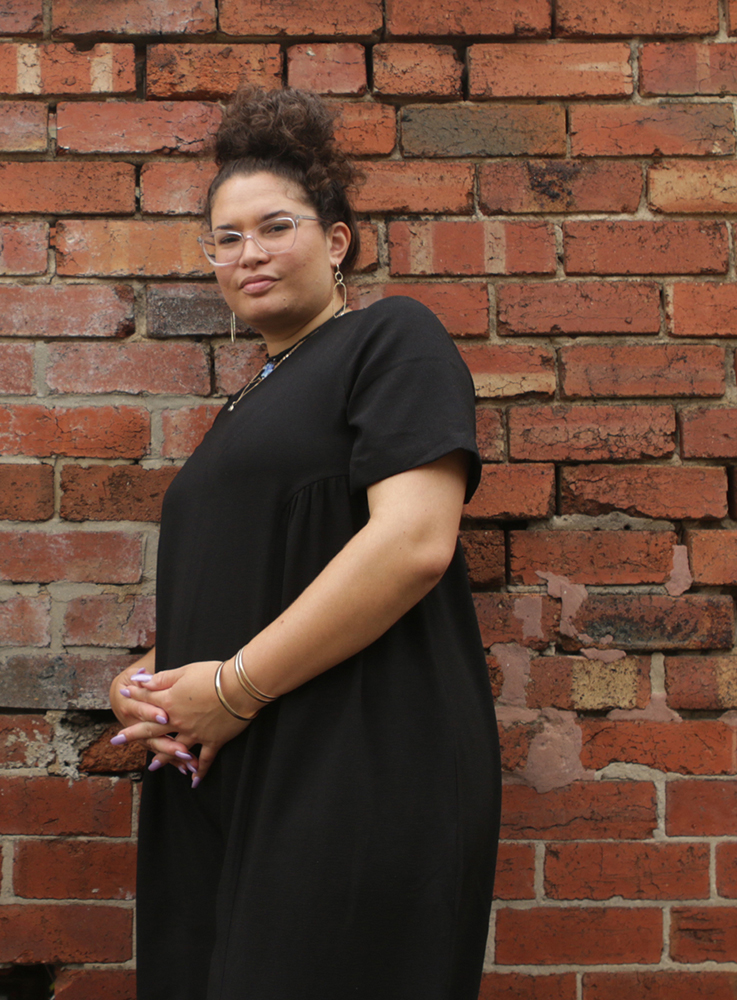 A light skinned African woman is leaning against a red brick wall, holding her hands together, wearing a black jumpsuit with short sleeves, with loose curly hair tied up high. She has clear framed square glasses and gives a small smile, while wearing fine gold earrings, some necklaces, and three gold bangles on one arm, and has lilac long nails.