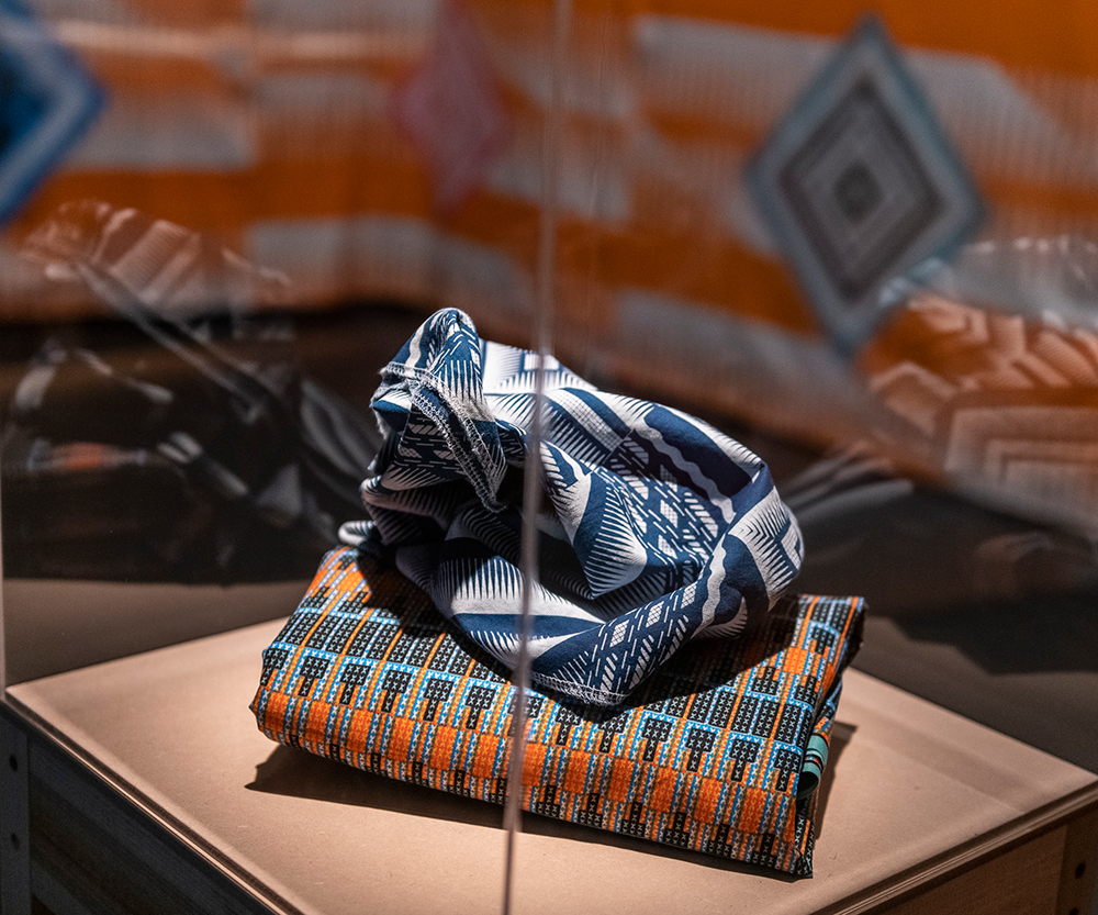 A white and blue fabric sits scrunched on top of an orange, teal, white and black patterned fabric folded neatly and is in focus. They sit on top of a square shaped MDF board, inside a perpex box that is creating faint reflections on either side and the corner of the box creates a line down the middle of the image. In the background is a larger orange pattern print that has diamond shapes in pink, blue, teal, white and black, and is not in clear focus.