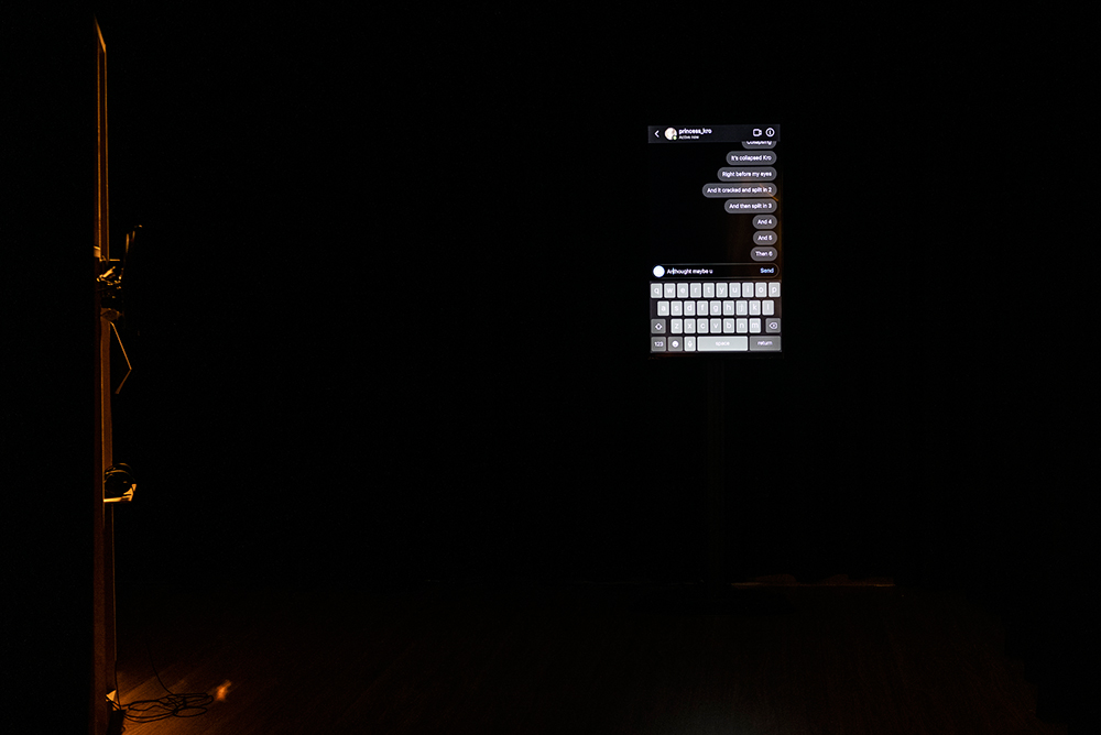 A screen in a dark room shows an instagram conversation, is opposite a low lit wood structure.