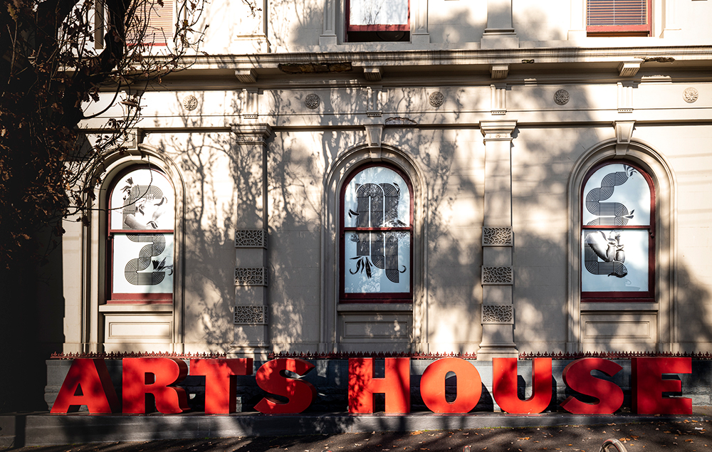 An old colonial building has a large shadow cast on it from a tree. Three arched windows have artworks with black on white designs that look like cornrows and braids that create the letters E, M, E. There are arms within the letters doing various gestures. Below the windows are large red letters that spell out Arts House.