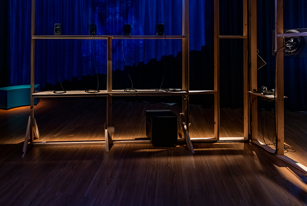 A dark room with floorboards and surrounding curtain has a timber structure spot lit in the middle of the room with speakkers and cords positioned on its shelves. There is a teal curved bench in the left background adjacent to a blue projection light that is shining on the back curtain.